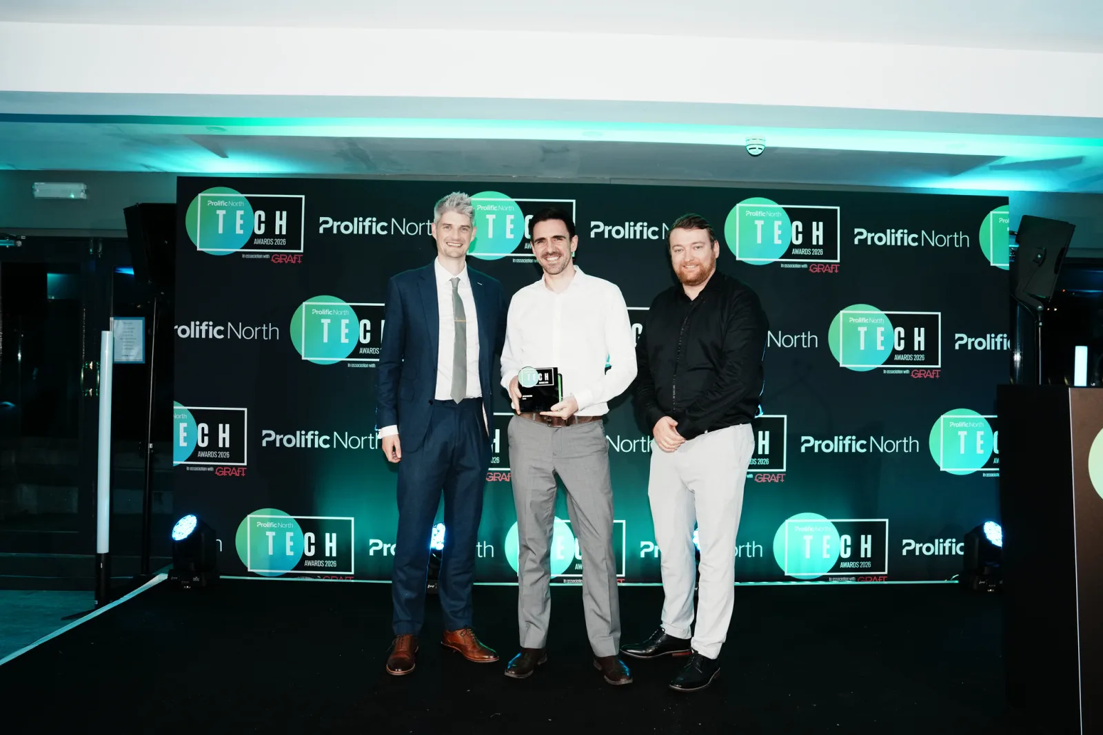 Nick, in a blue suit, and Mike, in a white shirt and holding the App of the year trophy, on stage with the award presenter in front of the Prolific North Tech Awards backdrop.